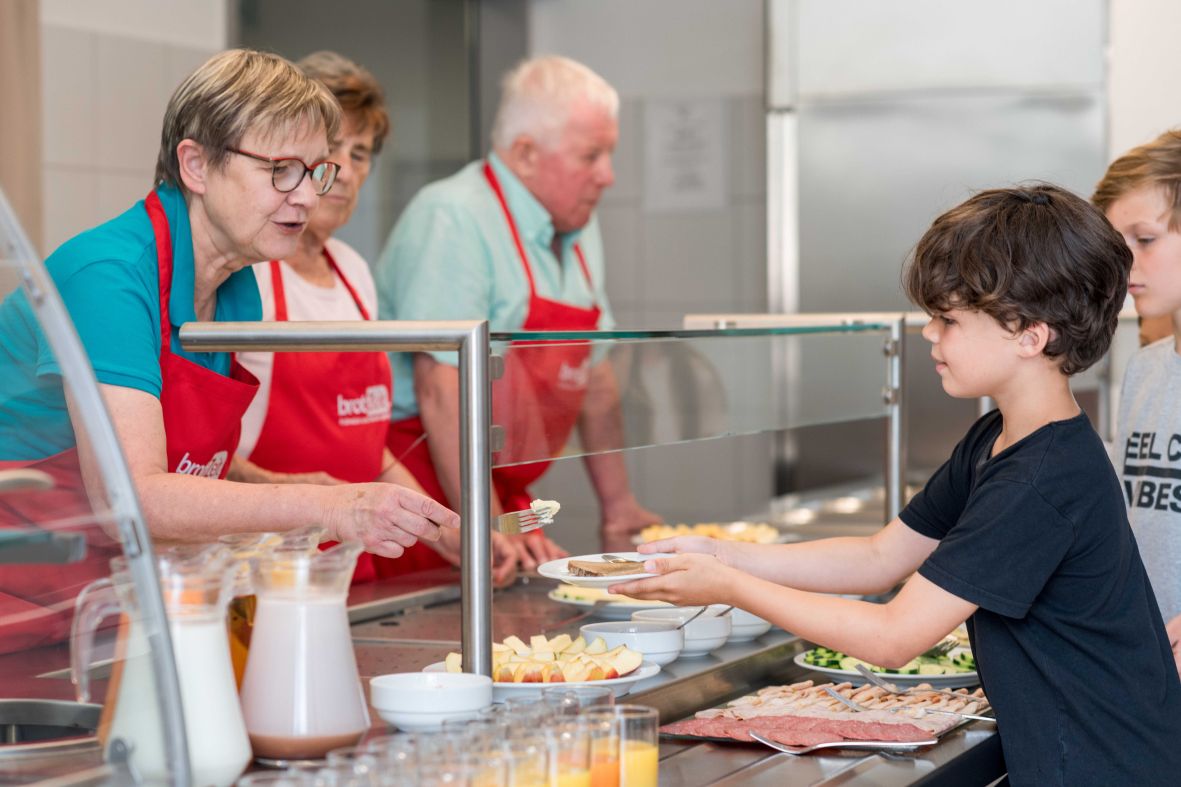 Kinder mit Teller bekommen Frühstück vonälterer Frau am Buffet gereicht